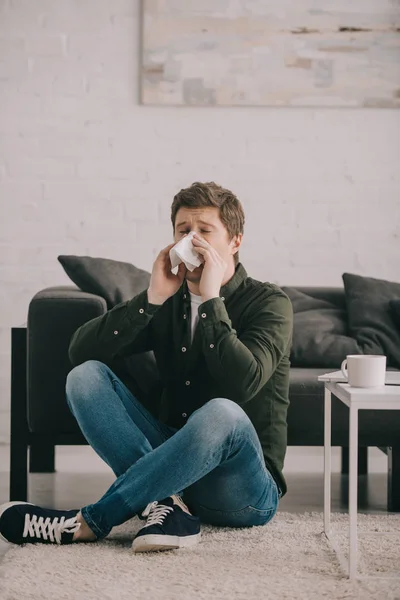 Mann sitzt mit gekreuzten Beinen auf Teppich und niest in Taschentuch neben Couchtisch mit Tasse Kaffee — Stockfoto