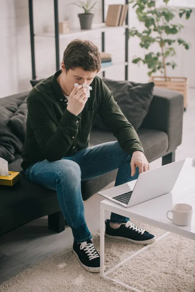Sick man sneezing in tissue while sitting on sofa and using laptop at home — Stock Photo