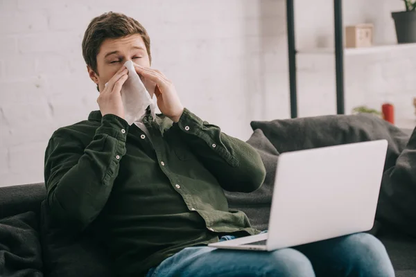 Homme malade ayant une allergie et des éternuements dans les tissus avec les yeux fermés tout en utilisant un ordinateur portable — Photo de stock