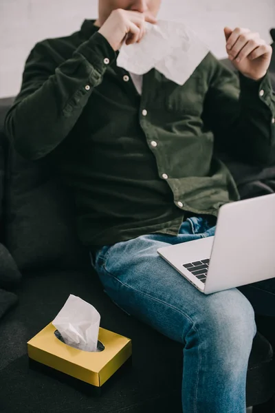 Cropped view of man having allergy and sneezing in tissue while using laptop — Stock Photo