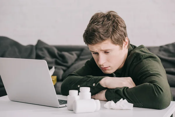 Upset man looking at bottles with pills on coffee table near laptop — Stock Photo