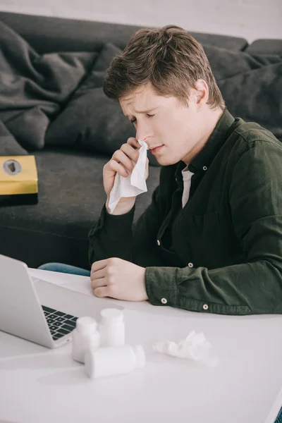 Засмучений чоловік тримає тканину, сидячи з закритими очима біля пляшок з таблетками і ноутбуком — стокове фото