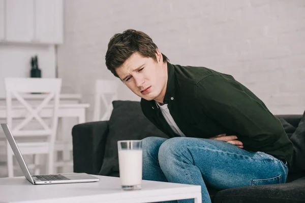 Расстроенный человек смотрит на стакан молока, держа желудок и сидя на диване — стоковое фото