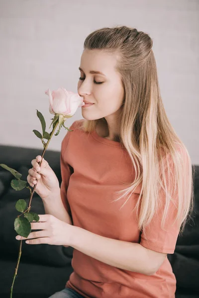 Fröhliches blondes Mädchen, das zu Hause mit geschlossenen Augen Rose riecht — Stockfoto