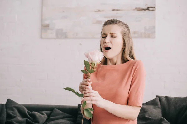 Attractive blonde girl with pollen allergy sneezing while holding rose at home — Stock Photo