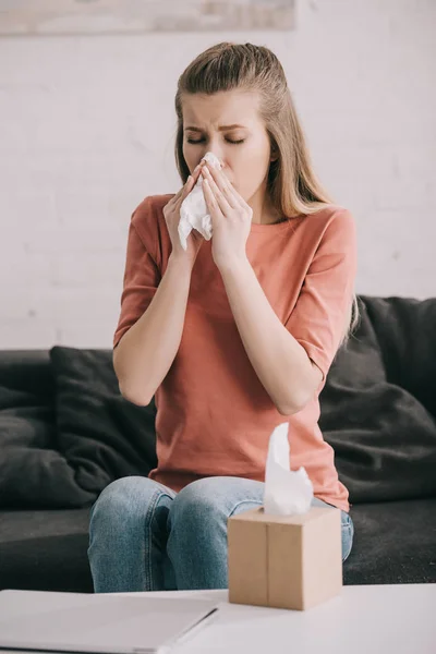 Attractive blonde girl sneezing while holding tissue near tissue box at home — Stock Photo