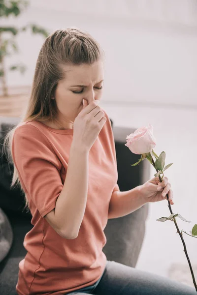 Привлекательная блондинка с аллергией на пыльцу чихает, держа цветок дома — стоковое фото