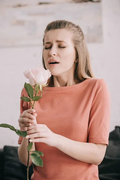 Hermosa chica rubia con alergia al polen estornudos con los ojos cerrados mientras sostiene rosa en casa - foto de stock