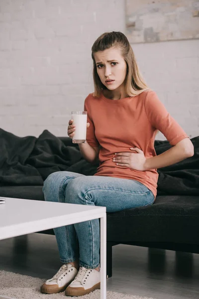 Aufgebrachte blonde Frau mit Laktoseintoleranz hält Glas Milch, während sie auf dem Sofa sitzt — Stockfoto