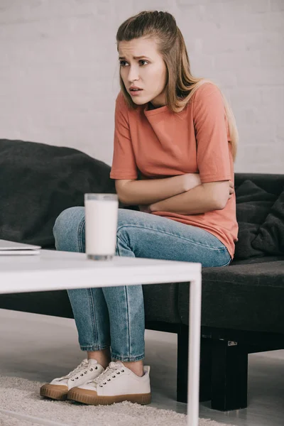 Mulher loira com intolerância à lactose segurando estômago perto de vidro de leite enquanto sentado no sofá — Fotografia de Stock