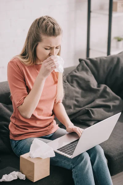 Attractive blonde woman sneezing in tissue while using laptop at home — Stock Photo