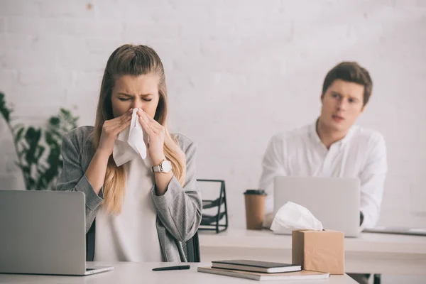 Blonde businesswoman sneezing in tissue near coworker in office — Stock Photo