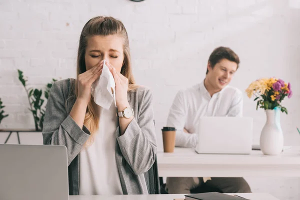 Blonde businesswoman sneezing in tissue with closed eyes near coworker in office — Stock Photo