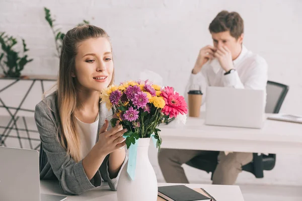 Selective focus of cheerful woman looking at flowers near coworker with pollen allergy — Stock Photo