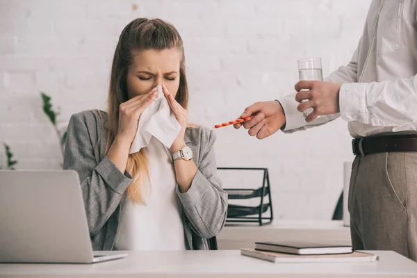 Блондинка-бизнесвумен чихает в ткани с закрытыми глазами рядом с коллегой с таблетками и стаканом воды — стоковое фото