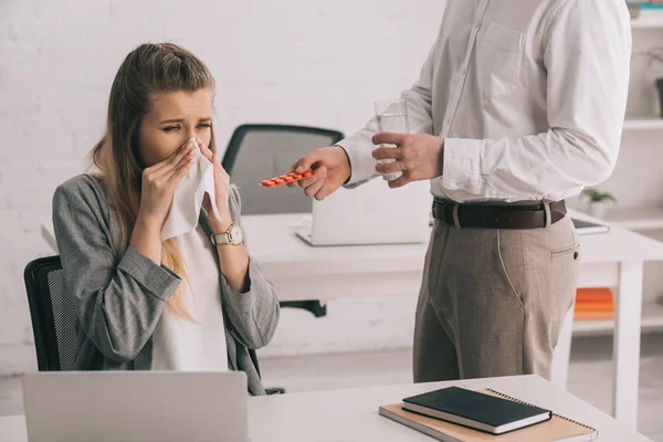 Cropped view of businessman holding pills and glass of water near blonde colleague sneezing in tissue — Stock Photo