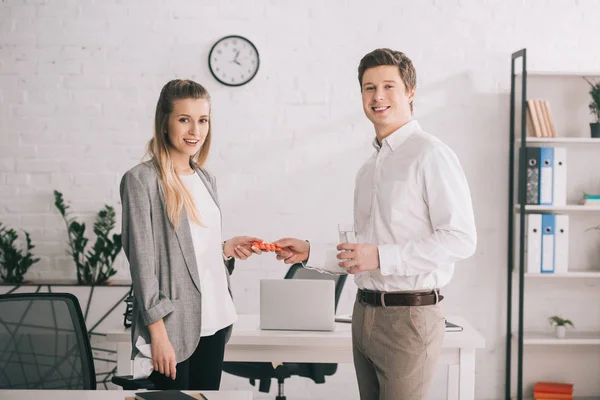 Happy businessman holding pills and glass of water near cheerful blonde colleague in office — Stock Photo