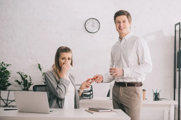 Happy businessman holding pills and glass of water near blonde colleague in office — Stock Photo