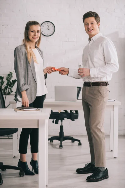 Cheerful businessman holding pills and glass of water near happy blonde colleague in office — Stock Photo