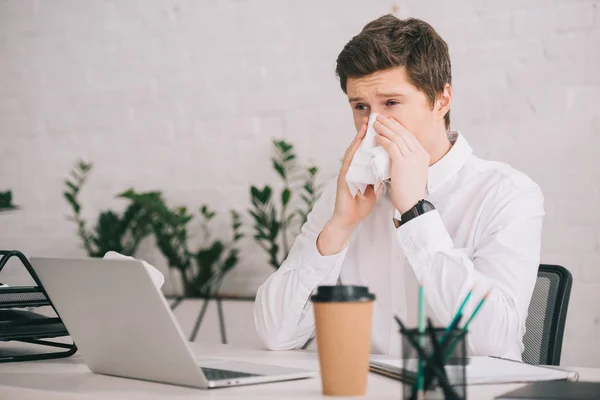 Schöner Geschäftsmann niest in Taschentuch, während er neben Laptop im Büro sitzt — Stockfoto