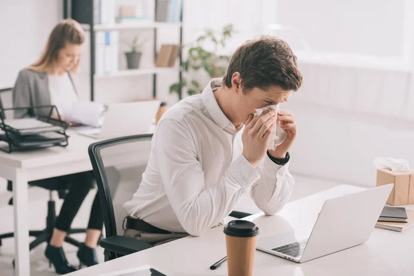 Enfoque selectivo de hombre de negocios estornudos en el tejido mientras mira el ordenador portátil cerca de la mujer de negocios en la oficina — Stock Photo