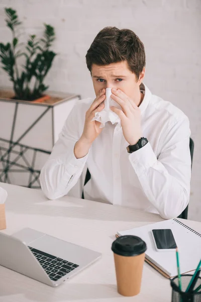 Guapo hombre de negocios estornudando en el tejido mientras está sentado cerca de la computadora portátil y el teléfono inteligente en la oficina - foto de stock