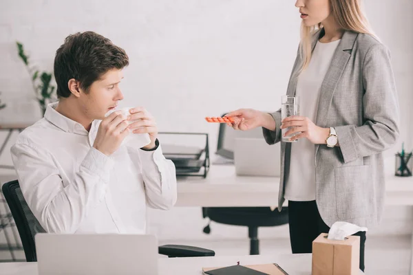 Cropped view of blonde woman holding pills and glass of water near coworker with tissue — Stock Photo