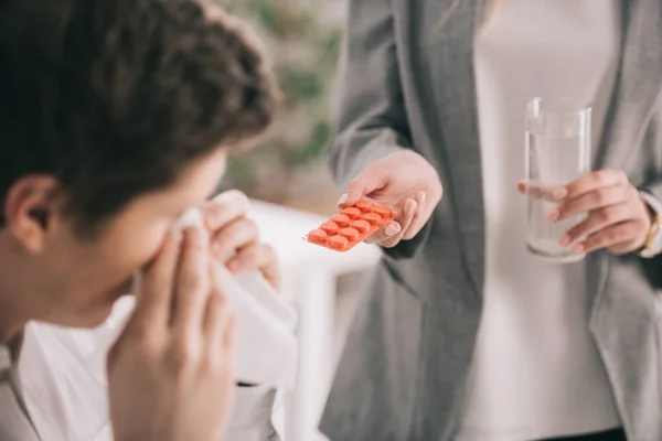 Vista recortada de la mujer sosteniendo píldoras y vaso de agua cerca de compañero de trabajo estornudando en tejido - foto de stock