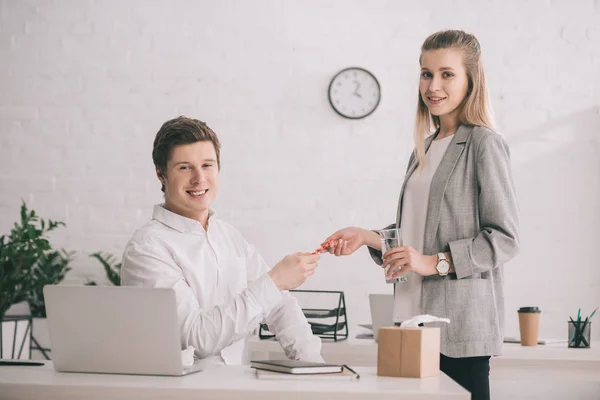 Cheerful businesswoman holding pills and glass of water near happy colleague in office — Stock Photo