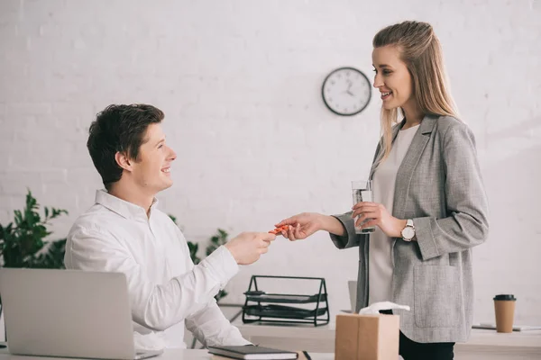 Alegre rubia empresaria sosteniendo píldoras y vaso de agua cerca feliz colega en la oficina - foto de stock