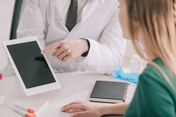 Vista ritagliata del medico in camice bianco che punta con il dito al tablet digitale con schermo bianco vicino al paziente biondo — Foto stock