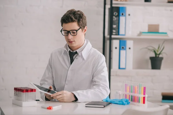 Gutaussehender Arzt mit Brille und weißem Mantel, der auf sein digitales Tablet blickt — Stockfoto