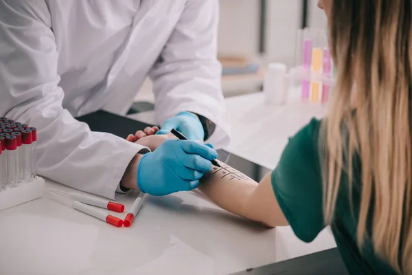 Ausgeschnittene Ansicht eines Arztes in weißem Mantel und Latexhandschuhen, der einen Filzstift in der Nähe der markierten weiblichen Hand hält — Stockfoto