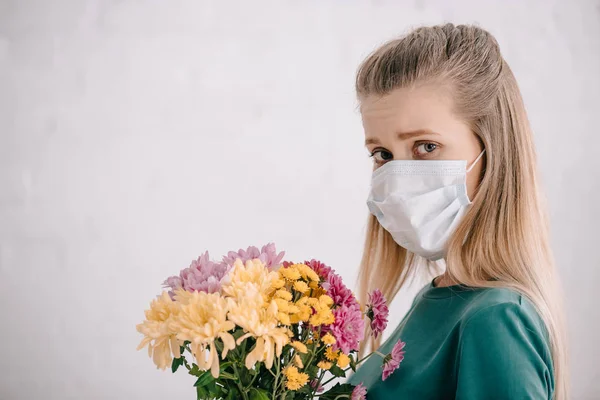 Mujer rubia con alergia al polen usando máscara médica y sosteniendo flores mientras mira la cámara - foto de stock