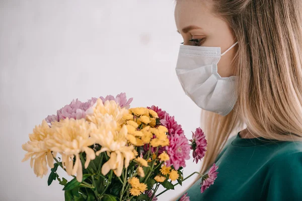 Blonde woman with pollen allergy wearing medical mask and looking at bouquet of flowers — Stock Photo