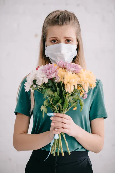 Femme blonde dans le masque médical tenant des fleurs dans les mains et regardant la caméra — Photo de stock