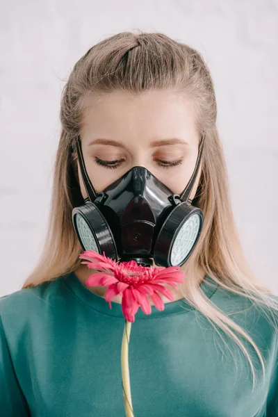 Donna bionda con allergia al polline con maschera respiratoria e guardando il fiore di gerbera rosa — Foto stock