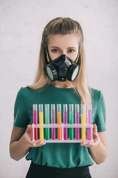 Mulher loira em máscara respiratória segurando tubos de ensaio coloridos — Fotografia de Stock