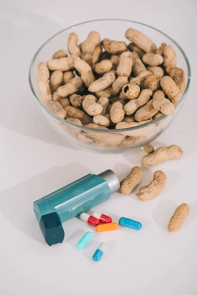 Glass bowl with nutritious peanuts near blue inhaler and pills on grey — Stock Photo