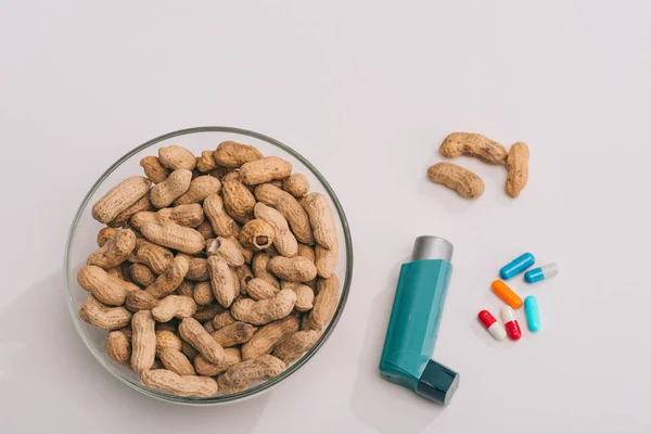 Vista superior de pílulas perto de inalador azul e tigela de vidro com amendoins saborosos em cinza — Fotografia de Stock