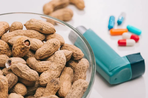 Foco selectivo de tazón de vidrio con sabrosos cacahuetes cerca de las píldoras y el inhalador azul en gris - foto de stock