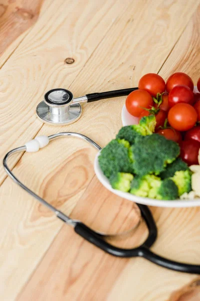 Вкусные органические овощи на тарелке возле стетоскопа на деревянной поверхности — стоковое фото