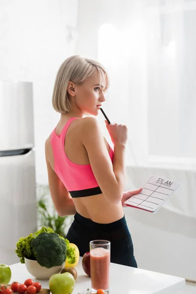 Mujer rubia pensativa en ropa deportiva con cuaderno con letras de plan en la cocina - foto de stock