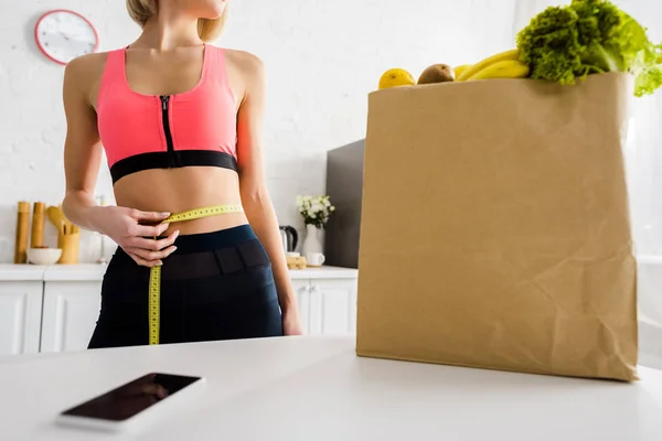 Vista recortada de la mujer sosteniendo cinta métrica en la cintura cerca de teléfono inteligente y bolsa de papel en la cocina - foto de stock