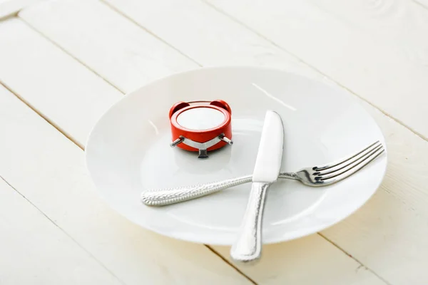 Белая тарелка со столовыми приборами и красными будильниками на деревянной поверхности — стоковое фото