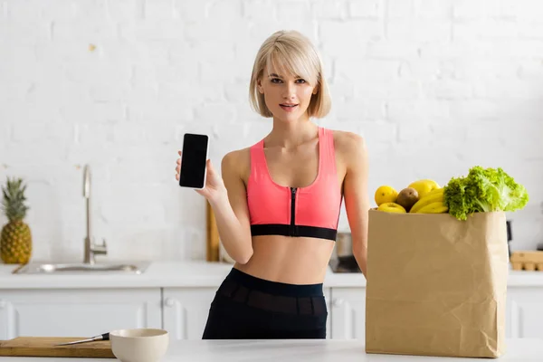 Mujer rubia en ropa deportiva sosteniendo smartphone con pantalla en blanco cerca de bolsa de papel con comestibles - foto de stock