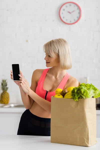 Весела блондинка в спортивному одязі тримає смартфон з порожнім екраном біля паперового мішка з продуктами — стокове фото