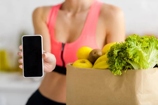 Обрезанный вид женщины в спортивной одежде держа смартфон с чистым экраном рядом с бумажным пакетом с продуктами — стоковое фото