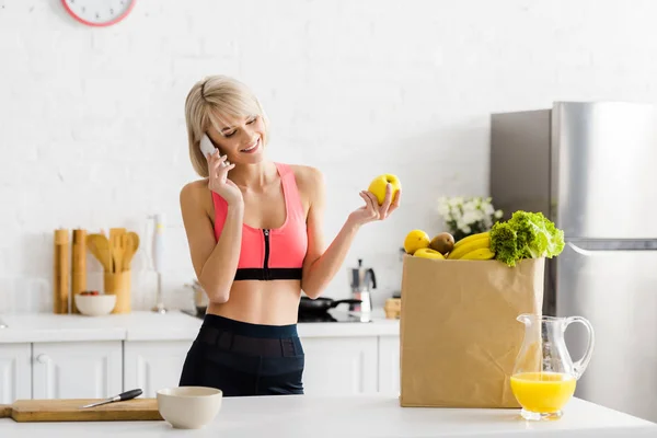 Happy blonde woman in sportswear talking on smartphone near paper bag with groceries — Stock Photo