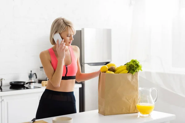 Attractive girl in sportswear talking on smartphone near paper bag with groceries — Stock Photo